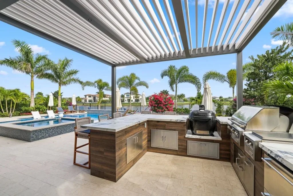 Outdoor kitchen under a louvered pergola beside the pool